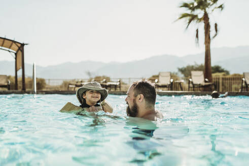 Vater und Kleinkind schwimmen im Pool mit Bergen und Palmen - CAVF91501