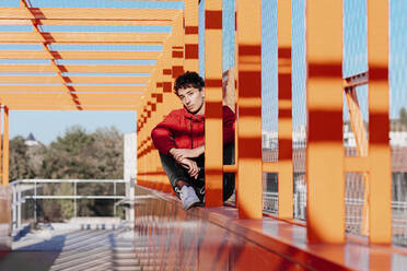 Ernster junger Mann sitzt auf orangefarbenem Brückengeländer - OGF00750