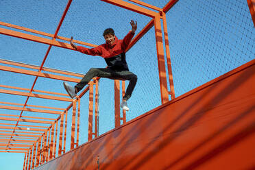Unbekümmerter junger Mann mit ausgestreckten Armen, der auf einer Brücke gegen einen klaren blauen Himmel springt - OGF00774