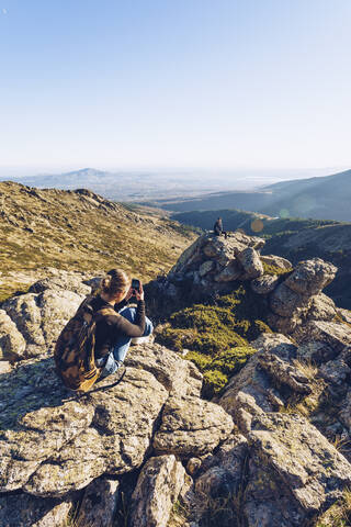 Young woman photographing male friend sitting on mountain against clear sky stock photo