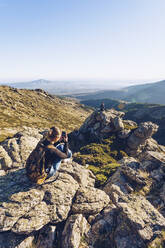 Young woman photographing male friend sitting on mountain against clear sky - RSGF00481