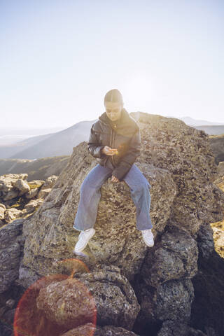 Wanderin, die ihr Smartphone benutzt, während sie auf einem Berg gegen den klaren Himmel sitzt, lizenzfreies Stockfoto