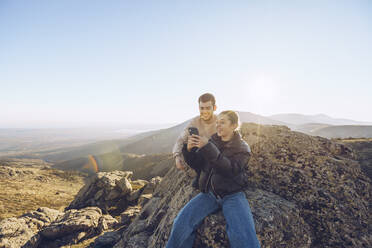 Friends using smart phone while sitting on mountain against clear sky - RSGF00487