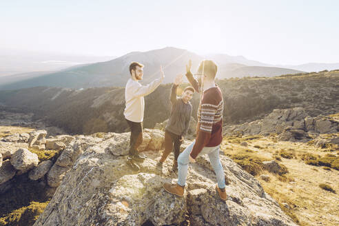 Cheerful friends giving high-five to each other while standing on mountain during sunny day - RSGF00492