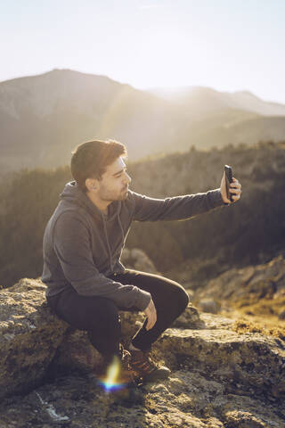 Young man taking selfie with smart phone while sitting on mountain during sunny day stock photo