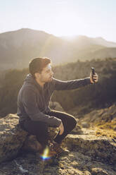 Junger Mann, der ein Selfie mit seinem Smartphone macht, während er an einem sonnigen Tag auf einem Berg sitzt - RSGF00494