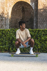 Lächelnder junger Mann, der auf einem Skateboard hockt und vor einem historischen Gebäude posiert - PNAF00510