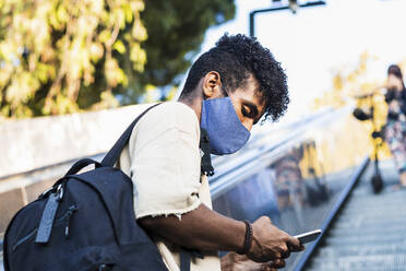 Young man wearing face mask using smart phone while standing on escalator - PNAF00512