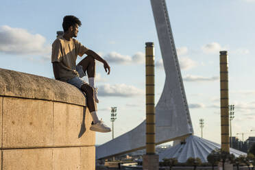Carefree young man sitting on top of retaining wall against sky in city during sunset - PNAF00513