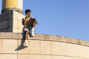 Young man talking over mobile phone while sitting on retaining wall against clear blue sky - PNAF00514
