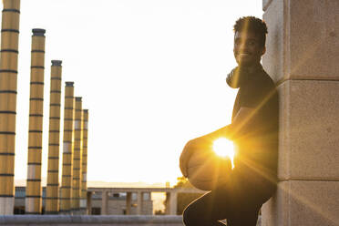 Sun shining through smiling man holding basketball while standing by wall at sunset - PNAF00527