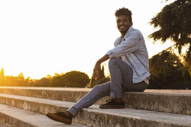 Smiling young man wearing denim jacket sitting on steps against clear sky at sunset - PNAF00528