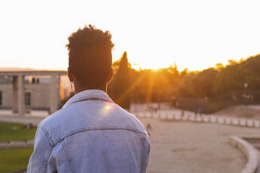 Junger Mann in Jeansjacke steht im Park bei Sonnenuntergang - PNAF00533