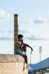 Junger Mann spricht über Smartphone, während er bei Sonnenuntergang auf einer Stützmauer sitzt - PNAF00537