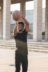 Lächelnder junger Mann spielt Basketball im Park bei Sonnenuntergang - PNAF00538