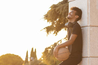 Fröhlicher junger Mann, der einen Basketball in der Hand hält, während er an der Wand steht, gegen einen klaren Himmel bei Sonnenuntergang - PNAF00540