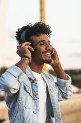 Close-up of smiling young man listening music through headphones against clear sky - PNAF00544