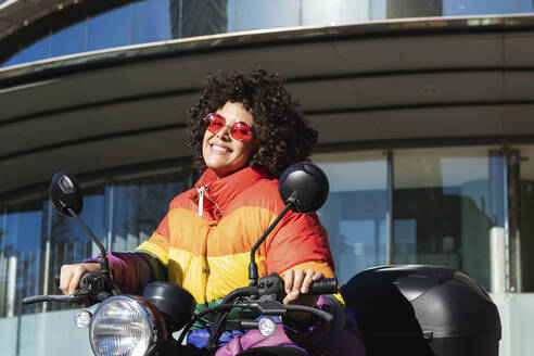 Frau mit Sonnenbrille und farbenfroher Jacke, die lächelnd auf einem Motorrad sitzt - PNAF00552