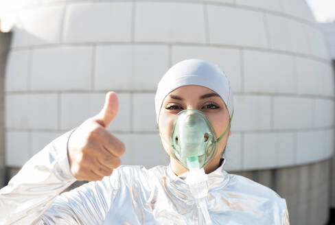 Woman wearing protective suit and oxygen mask showing thumbs up while standing against igloo - JCCMF00563