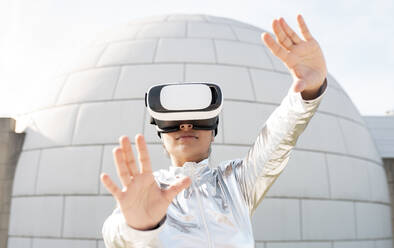 Woman wearing protective suit gesturing while using virtual reality headset against igloo - JCCMF00569