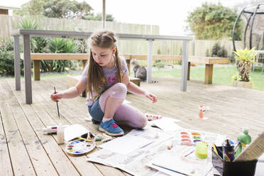 Junges Mädchen malt draußen auf einem Holzdeck mit einer Katze im Hintergrund - CAVF91515