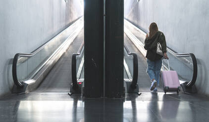 Young woman with suitcase walking towards moving walkway at railroad station - JAQF00133