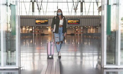 Young woman wearing face mask with suitcase walking at railroad station - JAQF00136