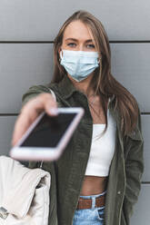 Young woman wearing face mask giving smart phone while standing against wall - JAQF00137