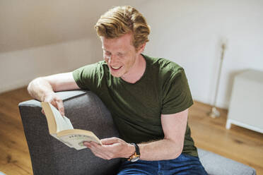 Lächelnder Mann liest ein Buch, während er auf der Couch im Wohnzimmer sitzt - MEF00061