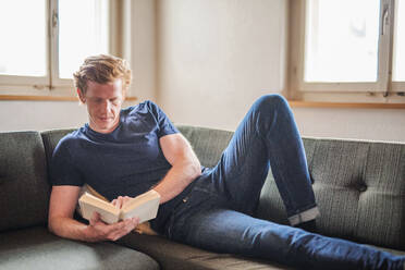 Man reading book while lying down on couch at home - MEF00063