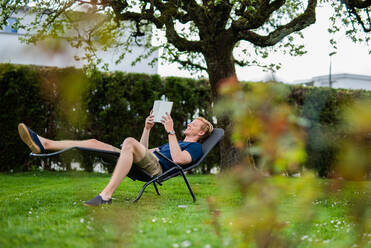 Happy man reading book while lying on chair in back yard - MEF00066