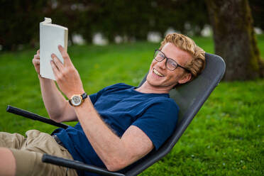 Mann lächelt und hält ein Buch auf einem Stuhl im Hof sitzend - MEF00067