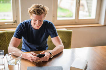 Lächelnder Mann, der sein Smartphone über einem Tisch zu Hause benutzt - MEF00077