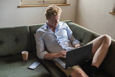 Männlicher Freiberufler arbeitet am Laptop, während er zu Hause auf der Couch sitzt - MEF00083