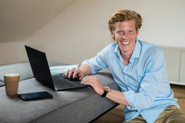 Smiling businessman with laptop by couch at home - MEF00091