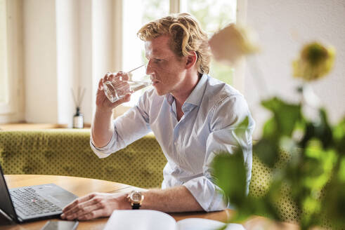 Männlicher Unternehmer trinkt Wasser, während er im Wohnzimmer am Laptop arbeitet - MEF00099