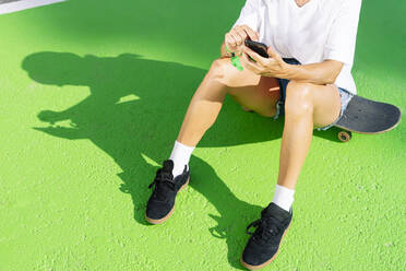 Low section of adult woman sitting on skateboard with smart phone in hands - PGF00372