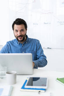 Portrait of smiling man using laptop in office - GIOF10464