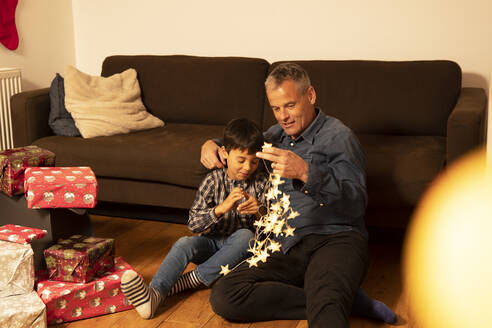 Vater und Sohn mit beleuchteter Weihnachtsbeleuchtung im Wohnzimmer sitzend - PMF01722