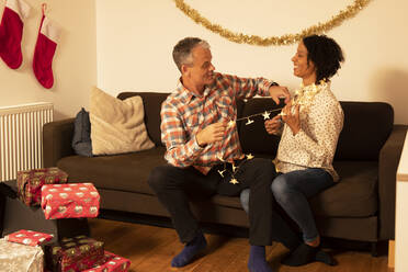 Cheerful couple playing with illuminated Christmas lights while sitting on sofa at home - PMF01724