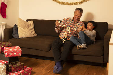 Vater und Sohn spielen mit Weihnachtsbeleuchtung, während sie zu Hause auf dem Sofa sitzen - PMF01725