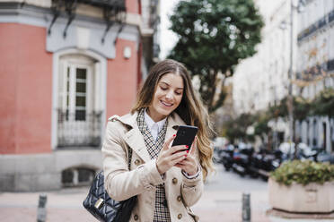 Happy woman carrying purse while using mobile phone in city - EBBF02057