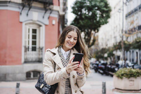 Glückliche Frau, die eine Handtasche trägt und ein Mobiltelefon in der Stadt benutzt - EBBF02057