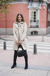 Smiling beautiful woman holding purse while standing on footpath in city - EBBF02058