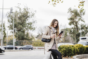 Fashionable woman with disposable coffee cup using smart phone while sitting on railing at street - EBBF02061