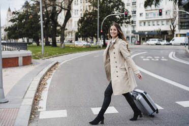 Cheerful woman with wheeled luggage crossing road in city - EBBF02064