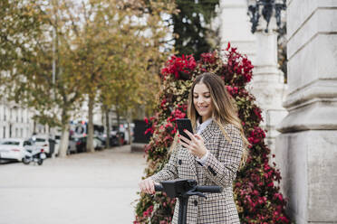 Lächelnde modische Frau mit Tretroller und Smartphone gegen Pflanze auf dem Bürgersteig - EBBF02069