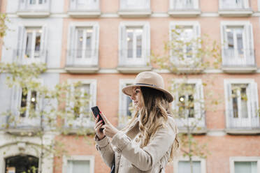 Mittlere erwachsene Frau mit Hut, die ein Mobiltelefon hält, während sie in der Stadt träumt - EBBF02074