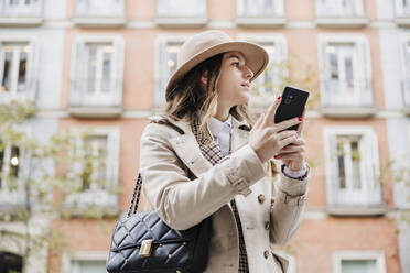 Fashionable woman looking away while holding smart phone against building in city - EBBF02075