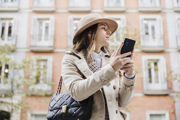 Modische Frau schaut weg, während sie ihr Smartphone gegen ein Gebäude in der Stadt hält - EBBF02075